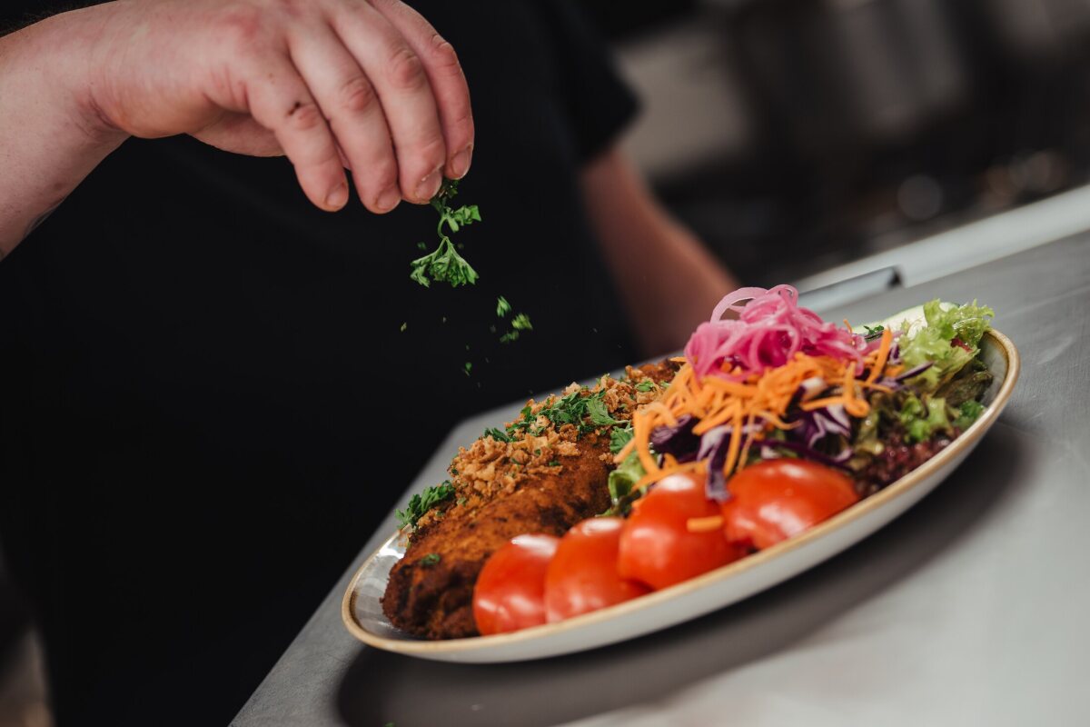Ein paniertes Gericht mit buntem Salat wird in der Küche mit frischen Kräutern bestreut.