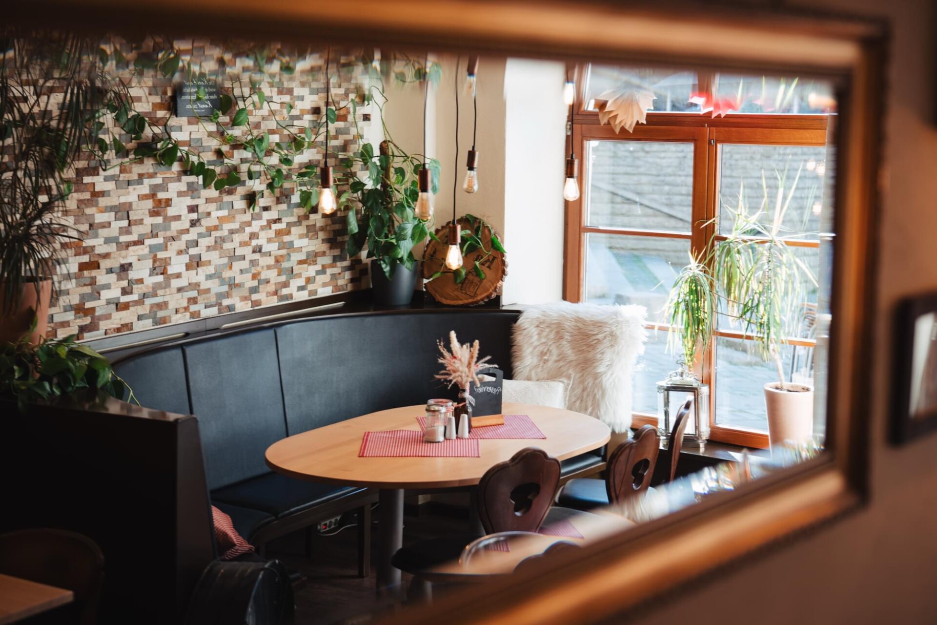 Blick durch einen Holzspiegel auf eine gemütliche Sitzecke mit rundem Tisch, Pflanzen und großem Fenster im Café Zeis.
