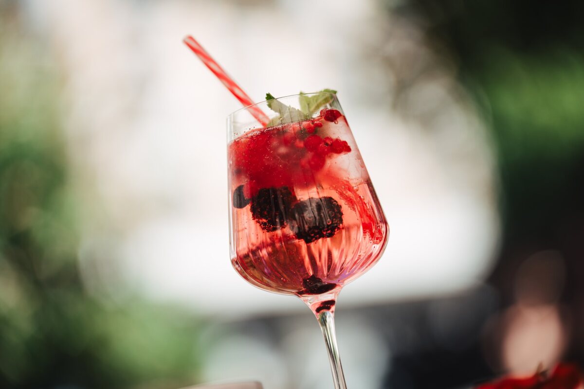 A glass of red berry drink with mint and a straw against a softly blurred green background.