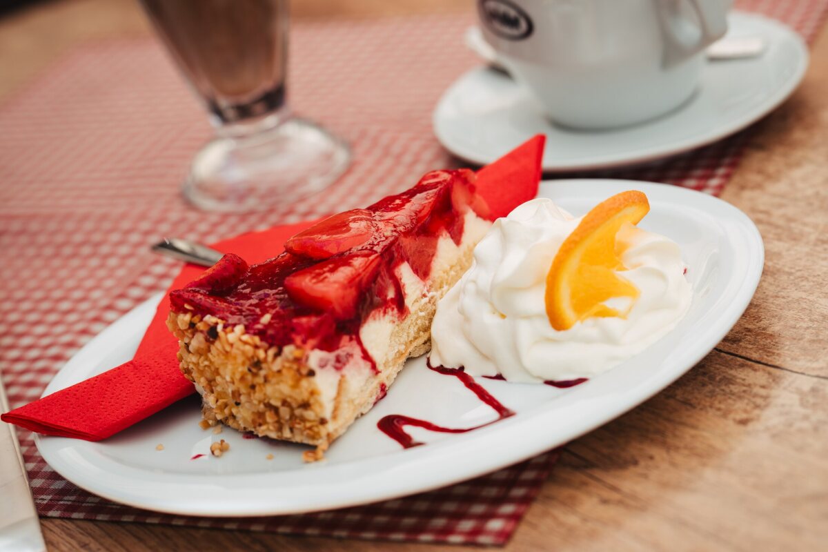 A slice of cake with red fruit and whipped cream, with coffee behind it on the checked tablecloth.